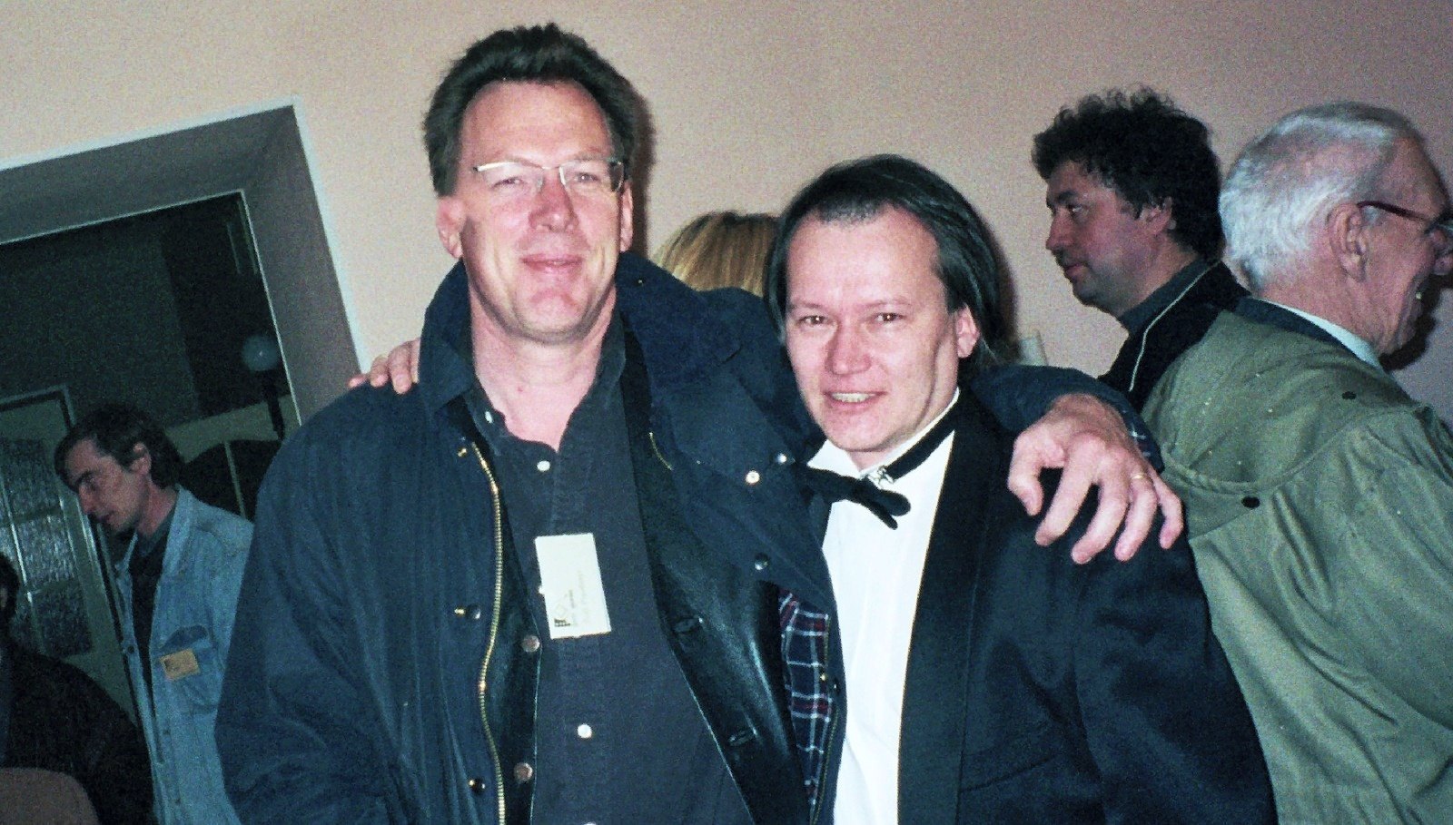 A group of men, one in a bow tie, posing arm in arm in a crowded room