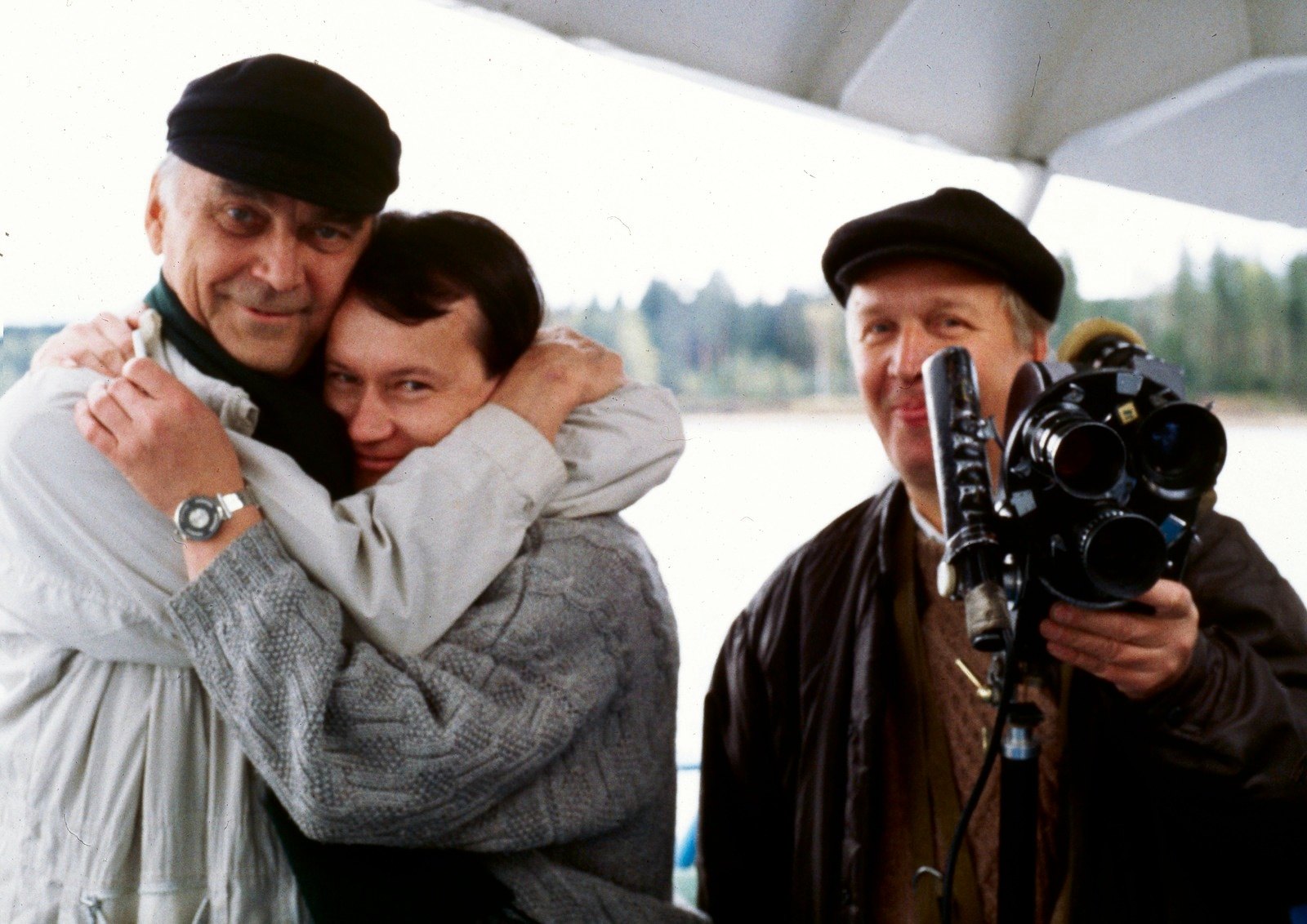 Two men embracing in the open air, a third man in a cap holding a film camera beside them