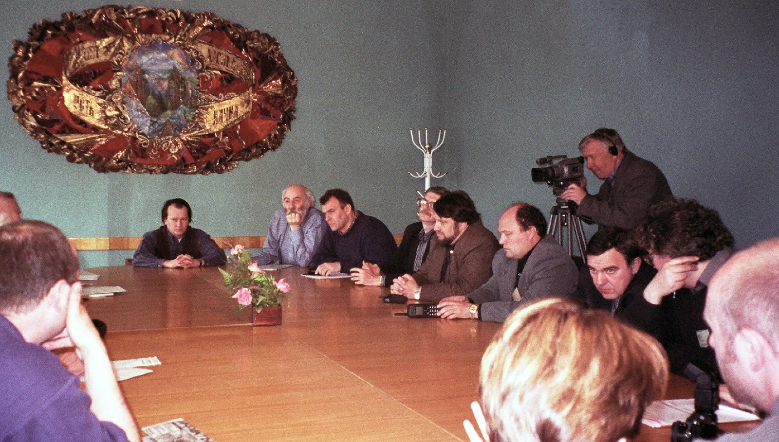 Men seated around a long table in a hall with a large ornamental emblem on the wall, a cameraman filming at the back