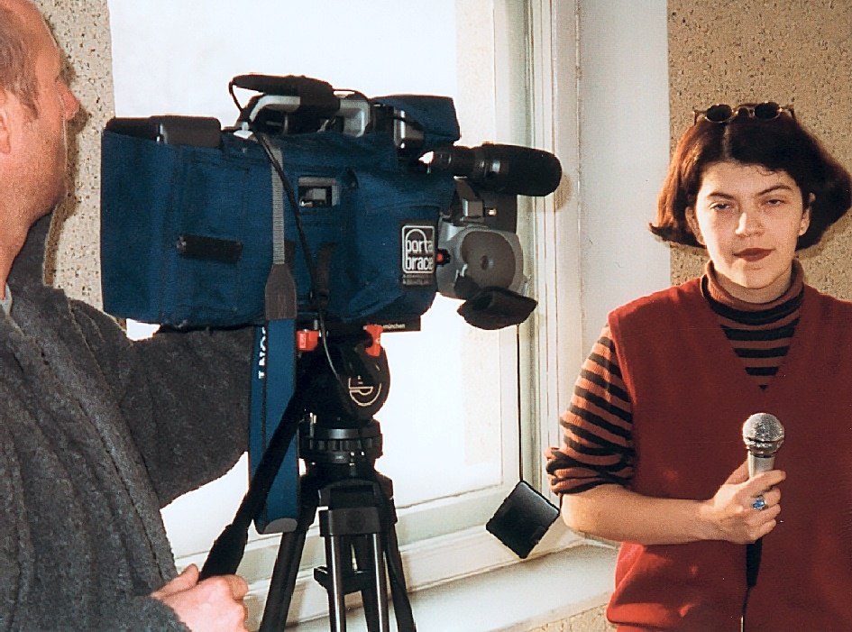 A young woman with a microphone stands by a window, facing a television camera on a tripod
