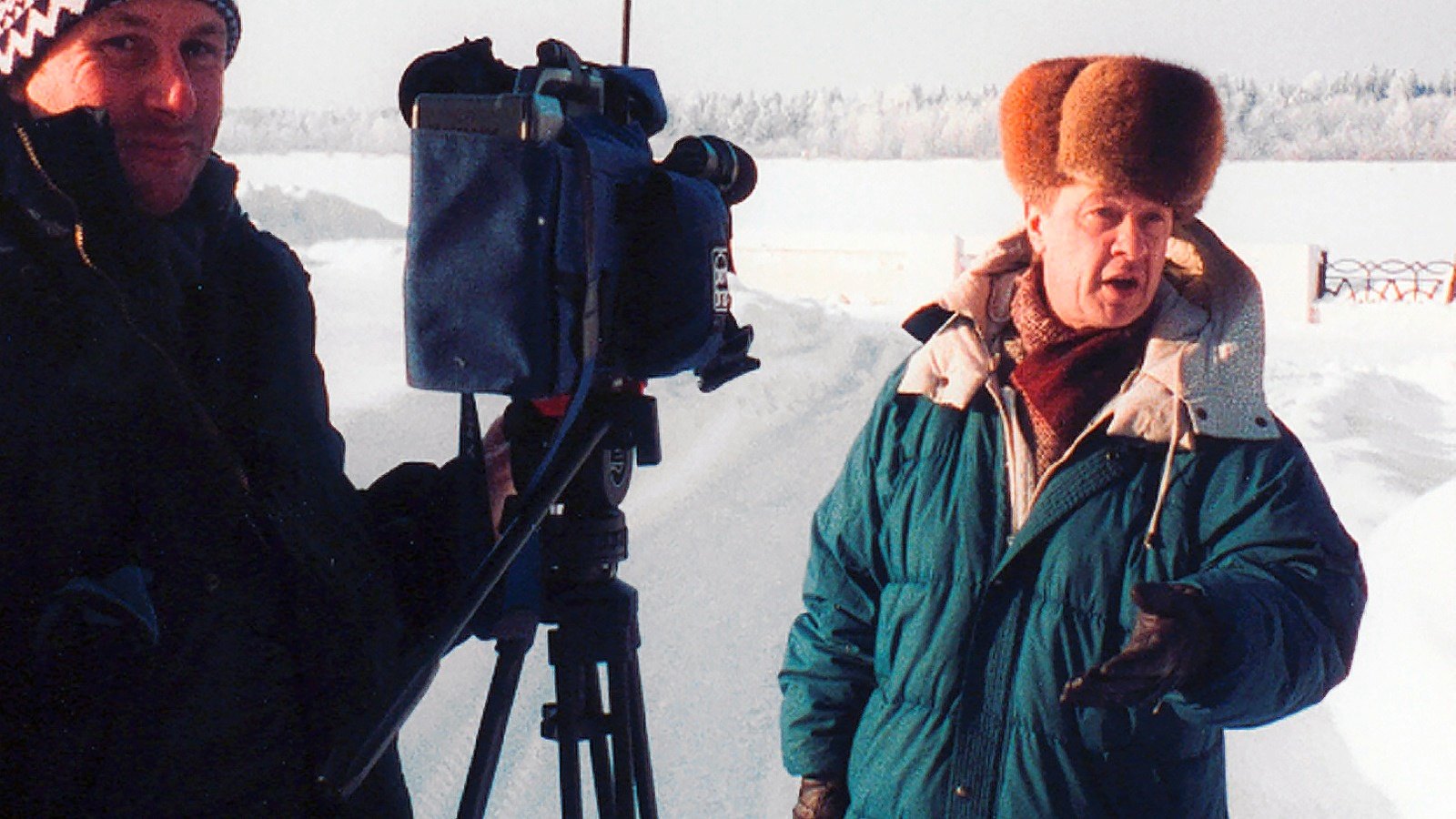 A cameraman behind a video camera on a tripod in deep snow, a man in a fur hat talking and gesturing
