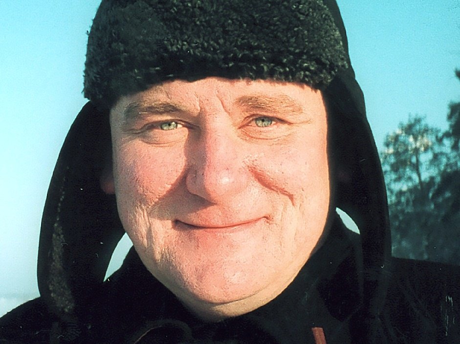 Close portrait of a smiling man in a dark fur-lined hood against a blue sky