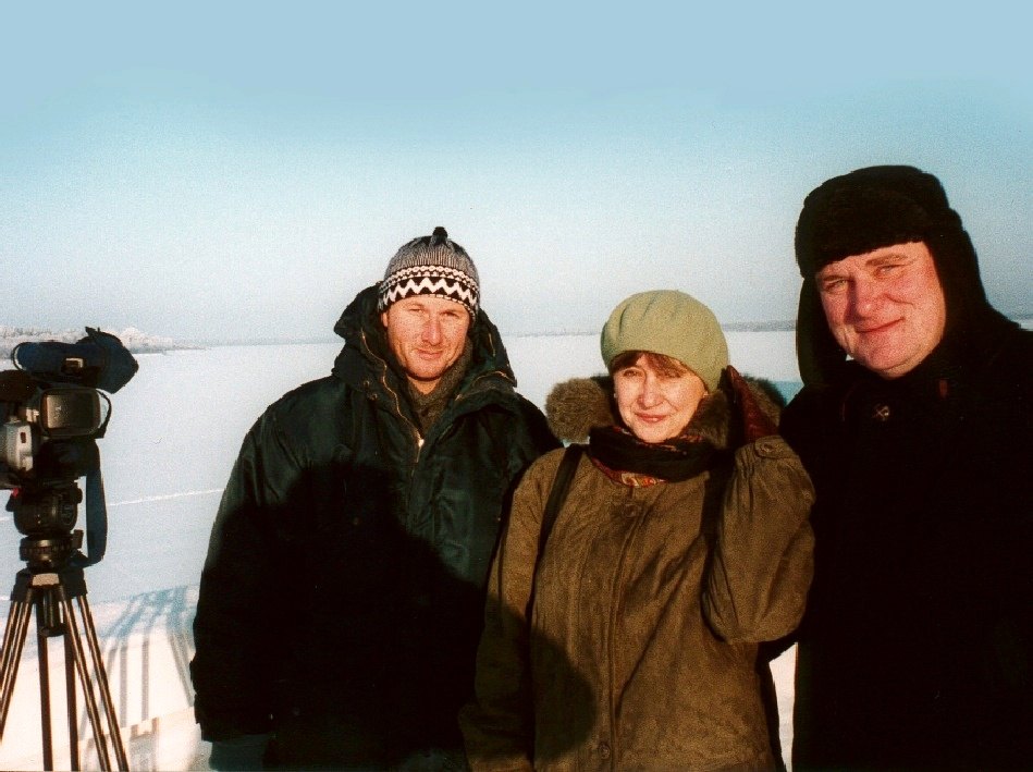 Two men and a woman in winter clothes side by side on a snowy plain, a video camera on a tripod at the left