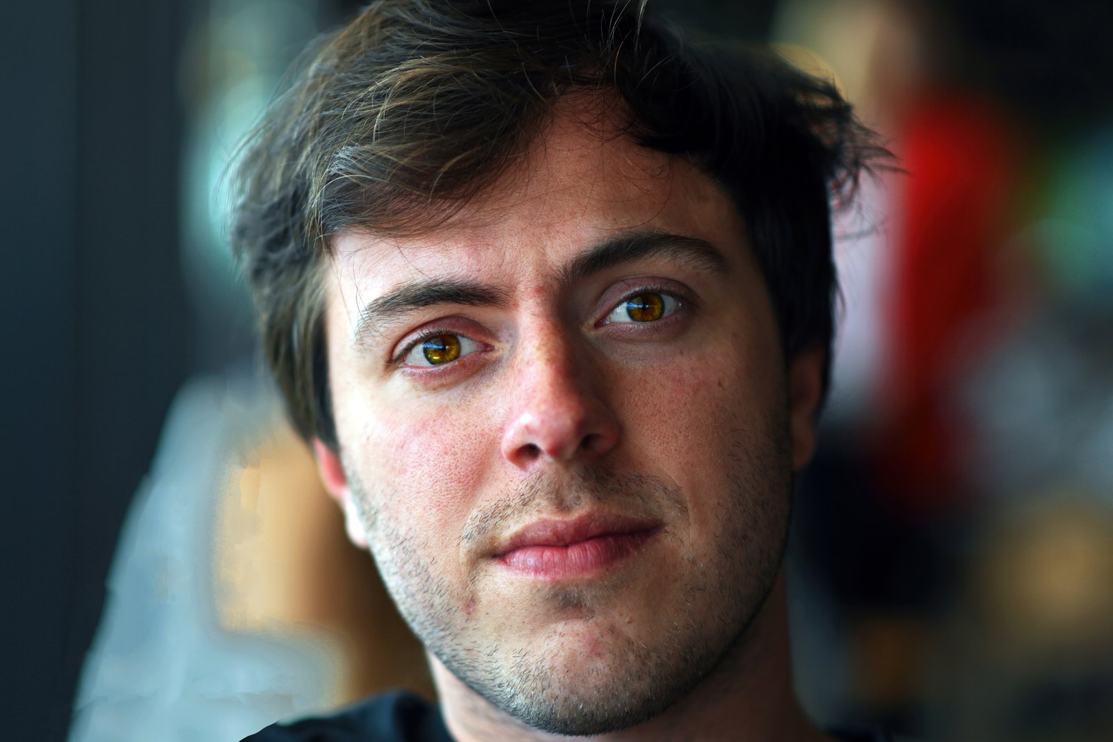 A young man with dark tousled hair looking into the camera, a blurred room behind him