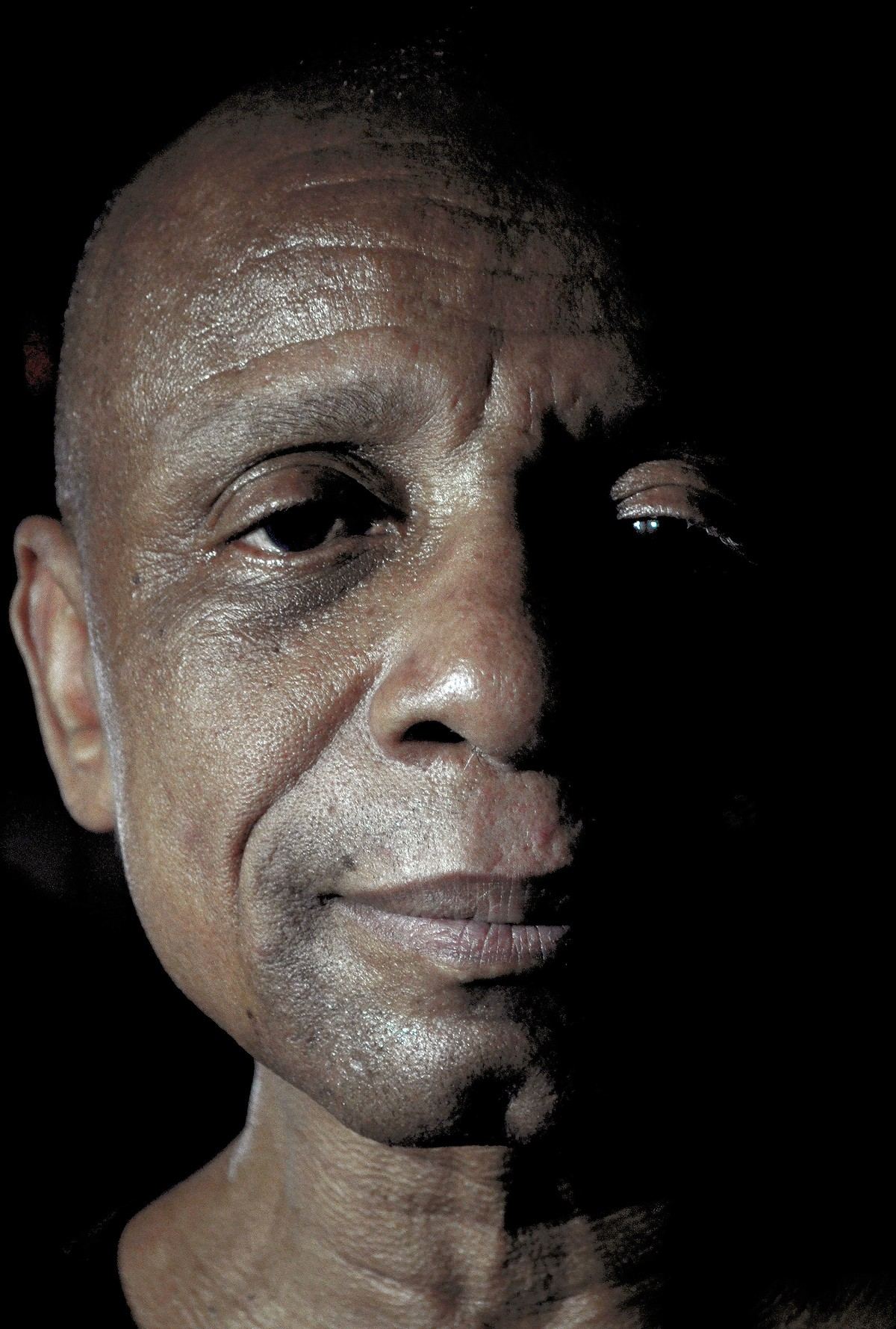 An older man with a shaved head, half his face in shadow, against a black background