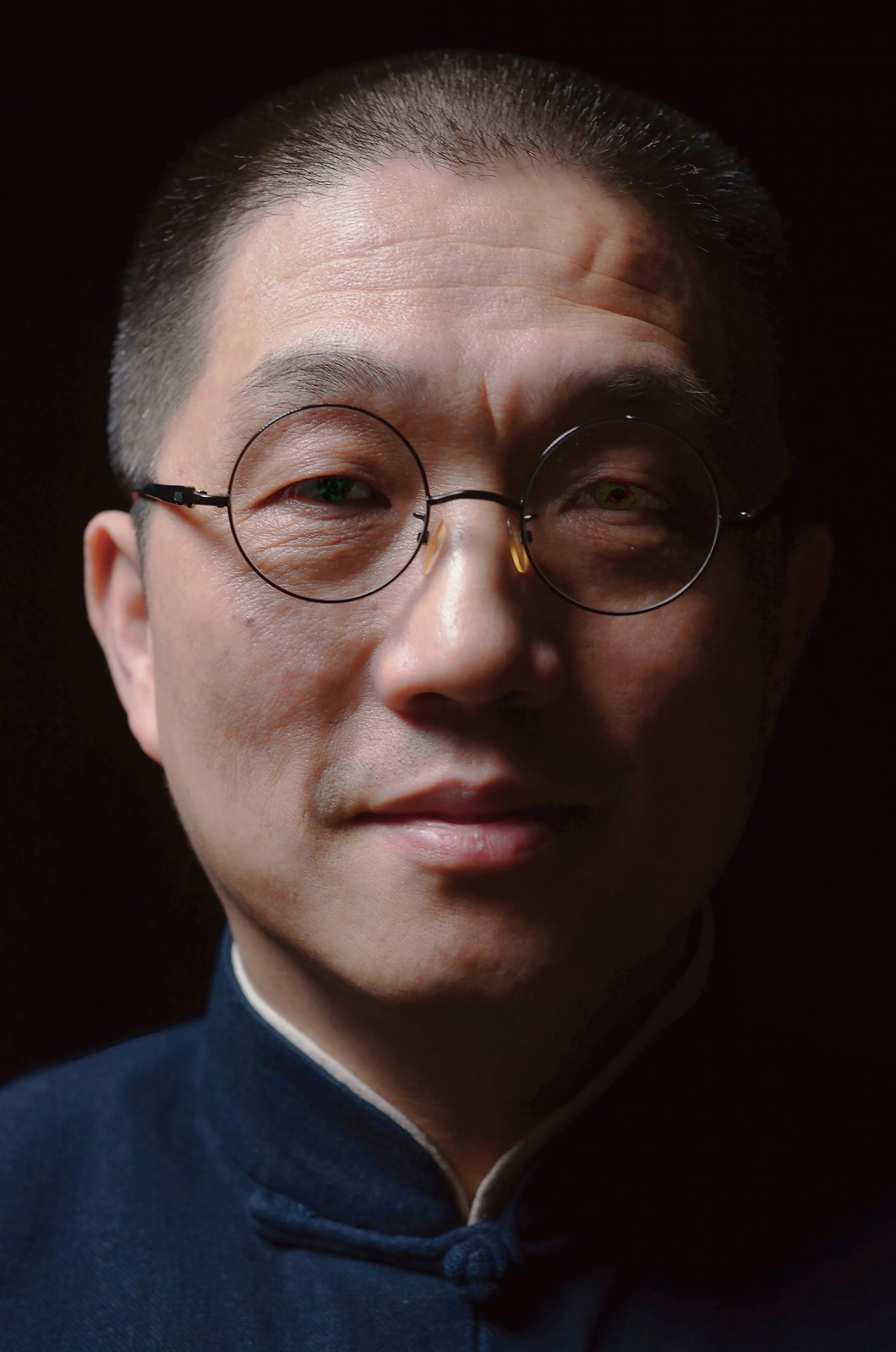 A man with round glasses and a dark high-collared jacket against a black background
