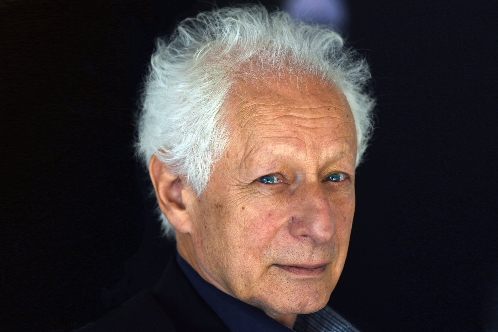 An older man with white curly hair and a dark jacket, against a dark background