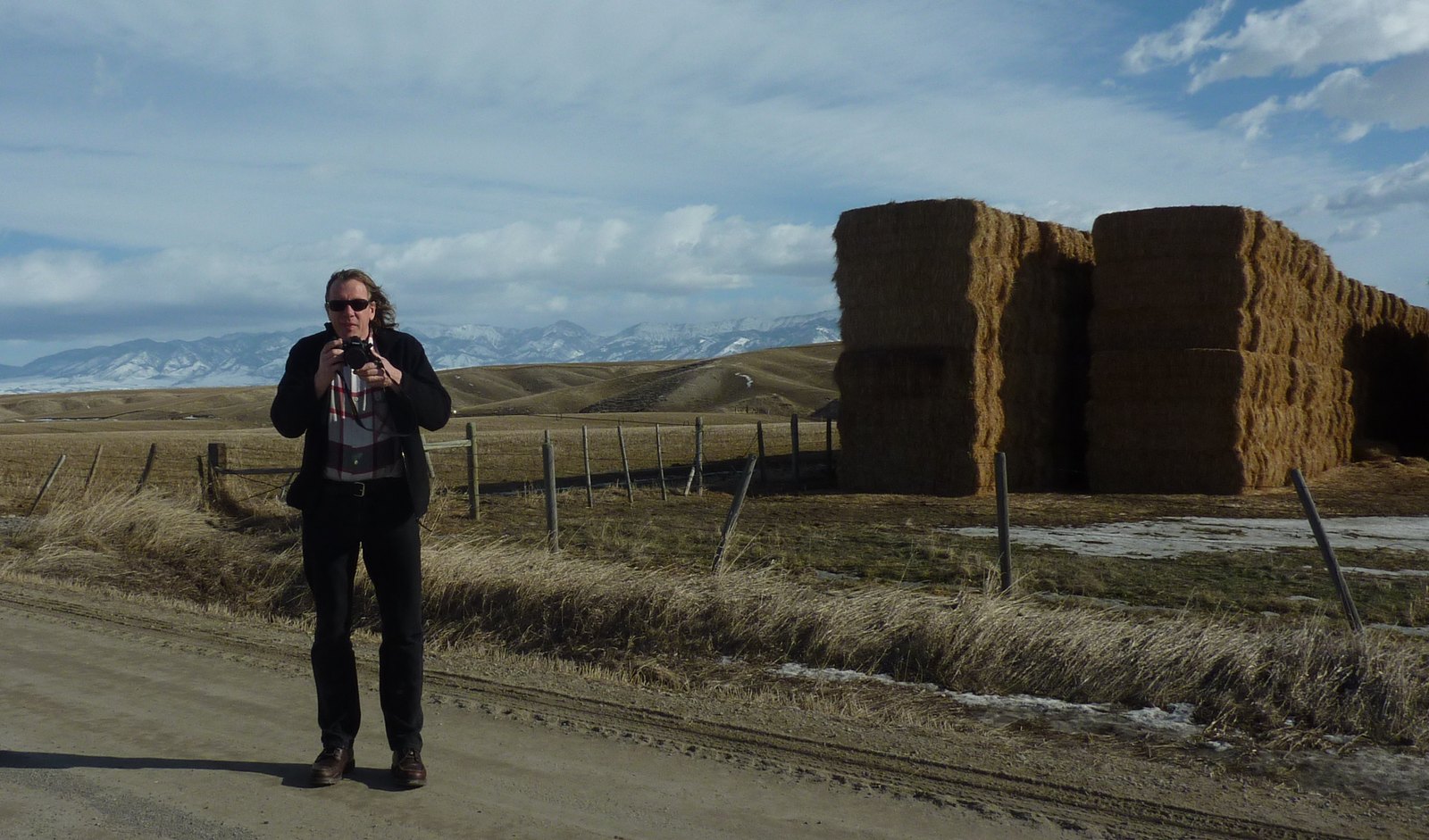 Rob Rombout on a gravel road in Montana, camera raised to his eye, with stacked hay bales and snowy mountains behind him